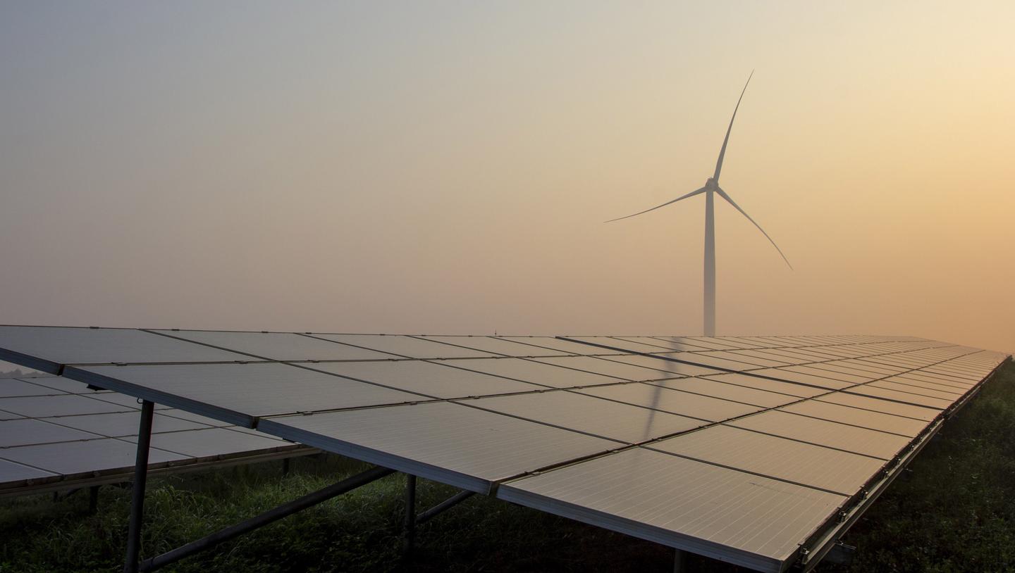 A photo representing renewable energy production, with solar panels in the foreground and a wind turbine in the background.